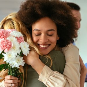 Women hug warmly holding pink and white flowers, with another person holding a champagne bottle in a cozy, indoor setting.