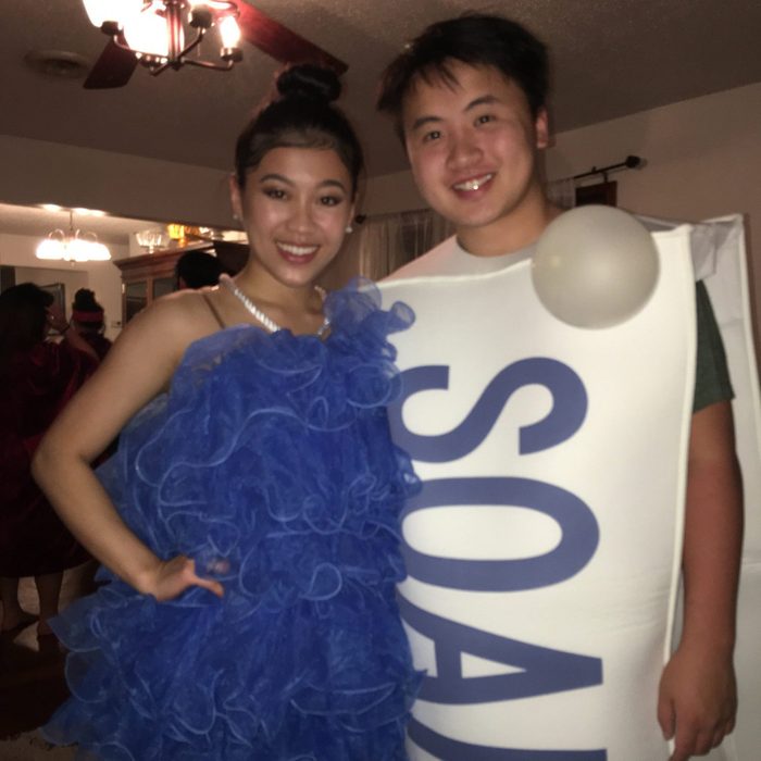 Two people are smiling; one in a blue ruffled dress, the other in a soap costume. They're in a warmly lit living room.