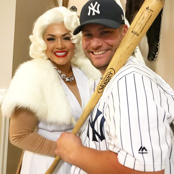 Man in Yankees jersey holds baseball bat; woman in white dress with fur stole smiles beside him. Indoor setting with subtle background details.