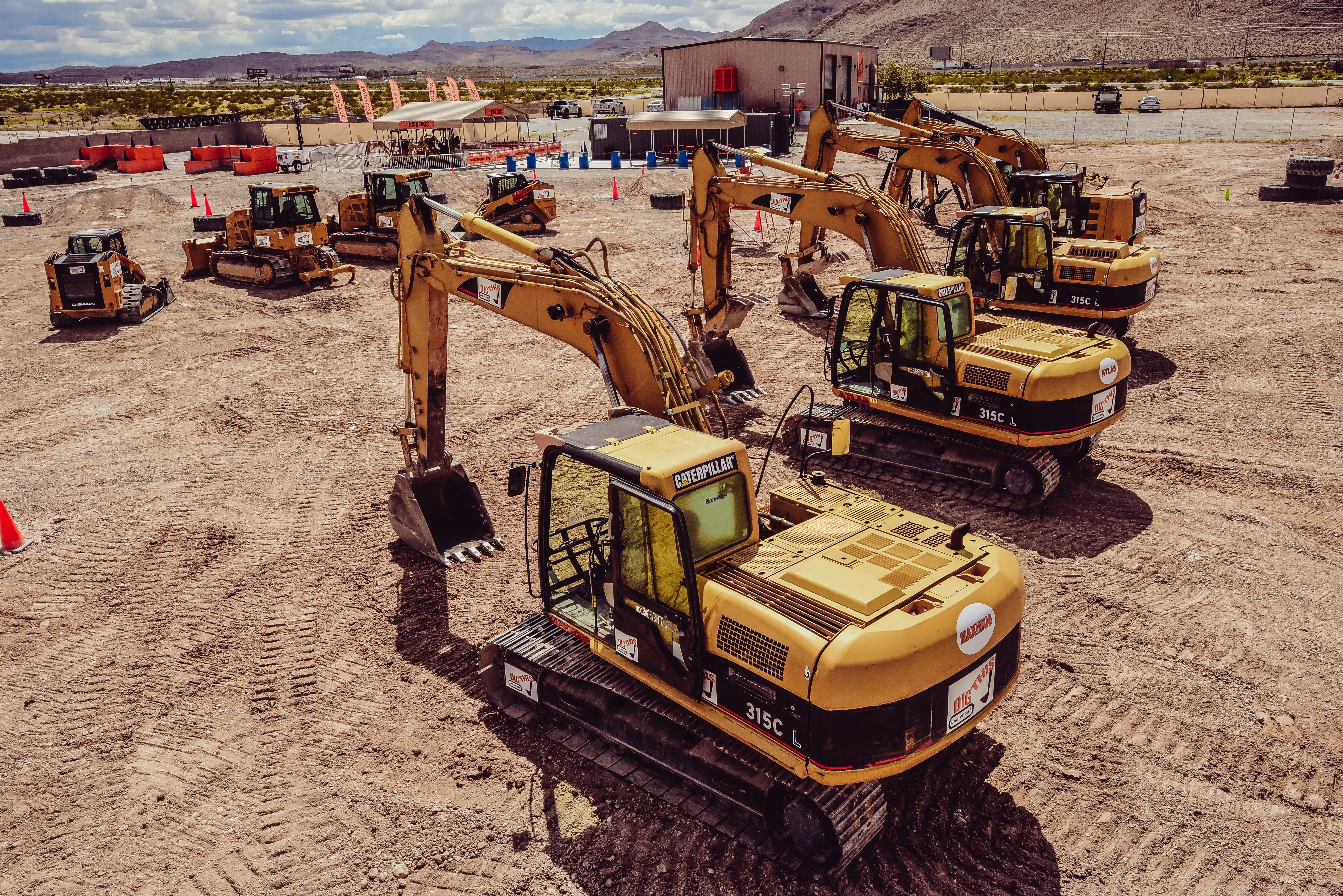 Excavators parked on dirt lot, surrounded by orange cones and machinery, mountains and buildings in the background. Text: "Caterpillar," "Maxim," "DIG THIS," "315C L."