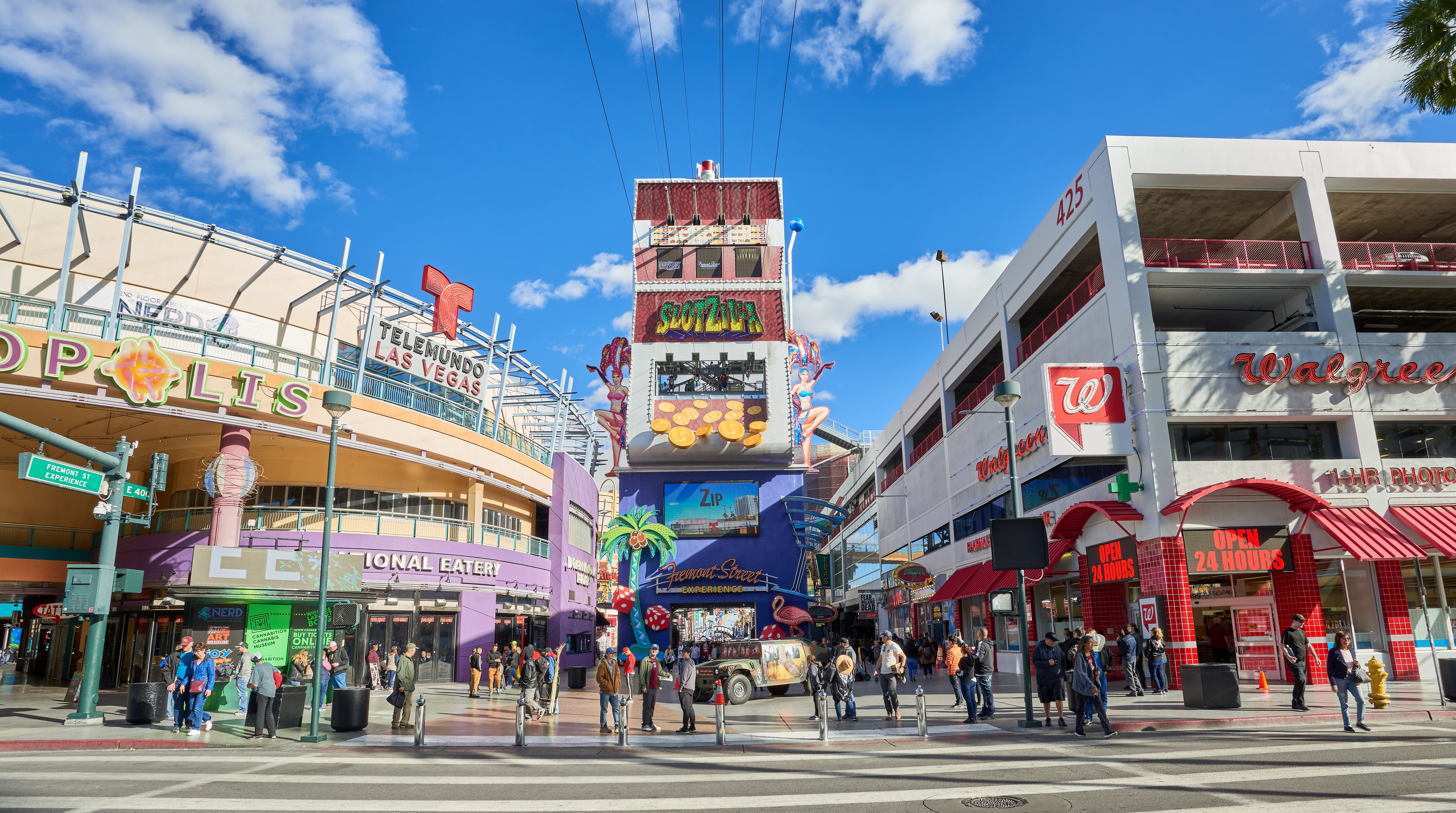 A bustling city street features crowds walking, flanked by colorful shops and a prominent SlotZilla zipline sign under a bright blue sky.