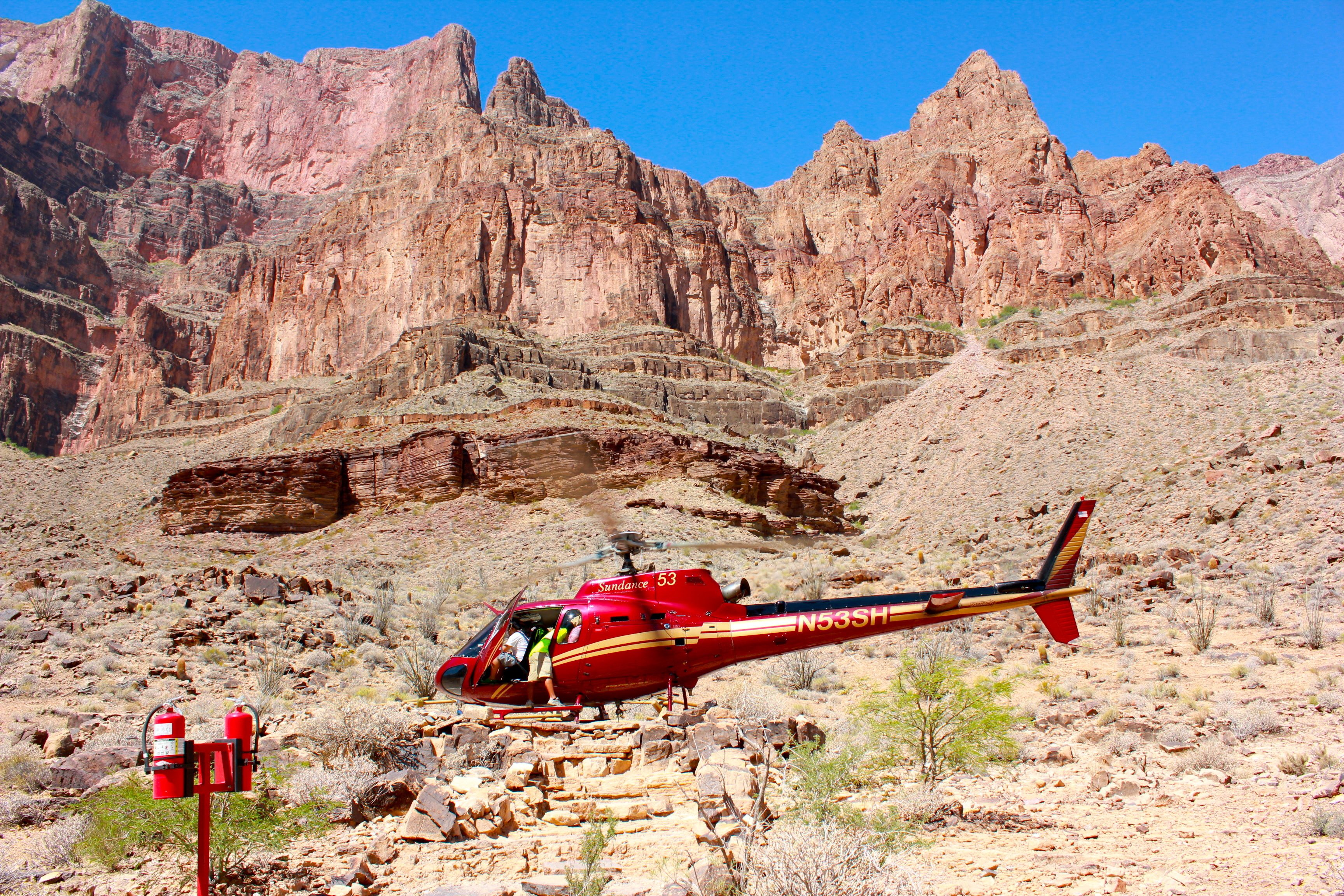 Helicopter hovering above rocky desert terrain with towering cliffs and clear blue sky. Red fire hydrant visible nearby. Tail number: N53SH.