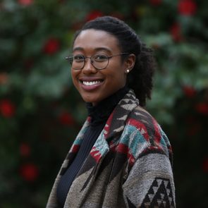 Person smiling, wearing glasses and patterned coat, standing outdoors; background is blurred with green foliage and red flowers.