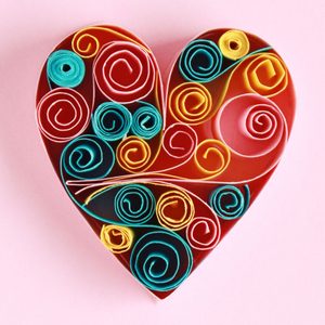 Colorful paper spirals form a heart shape on a pink background, creating an intricate, decorative pattern.