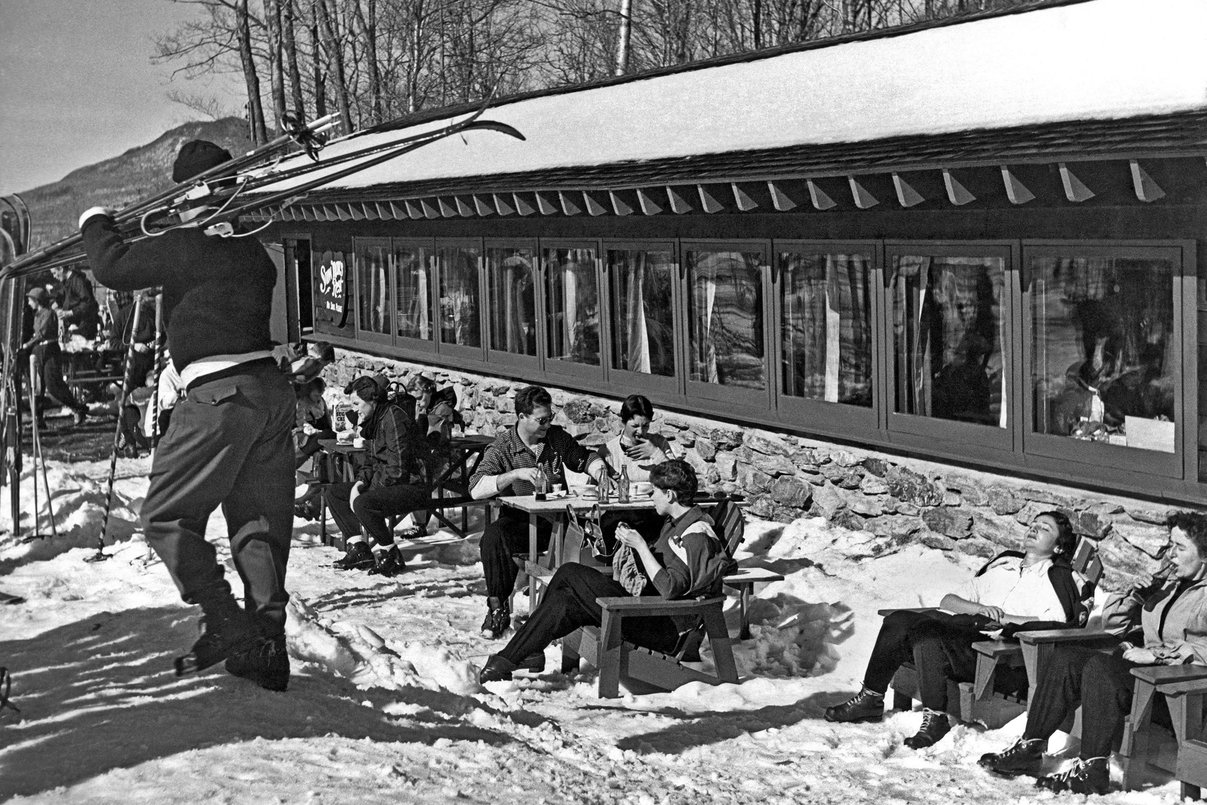 Skier carries skis past people dining and relaxing outside a snow-covered ski lodge, surrounded by trees and mountains.