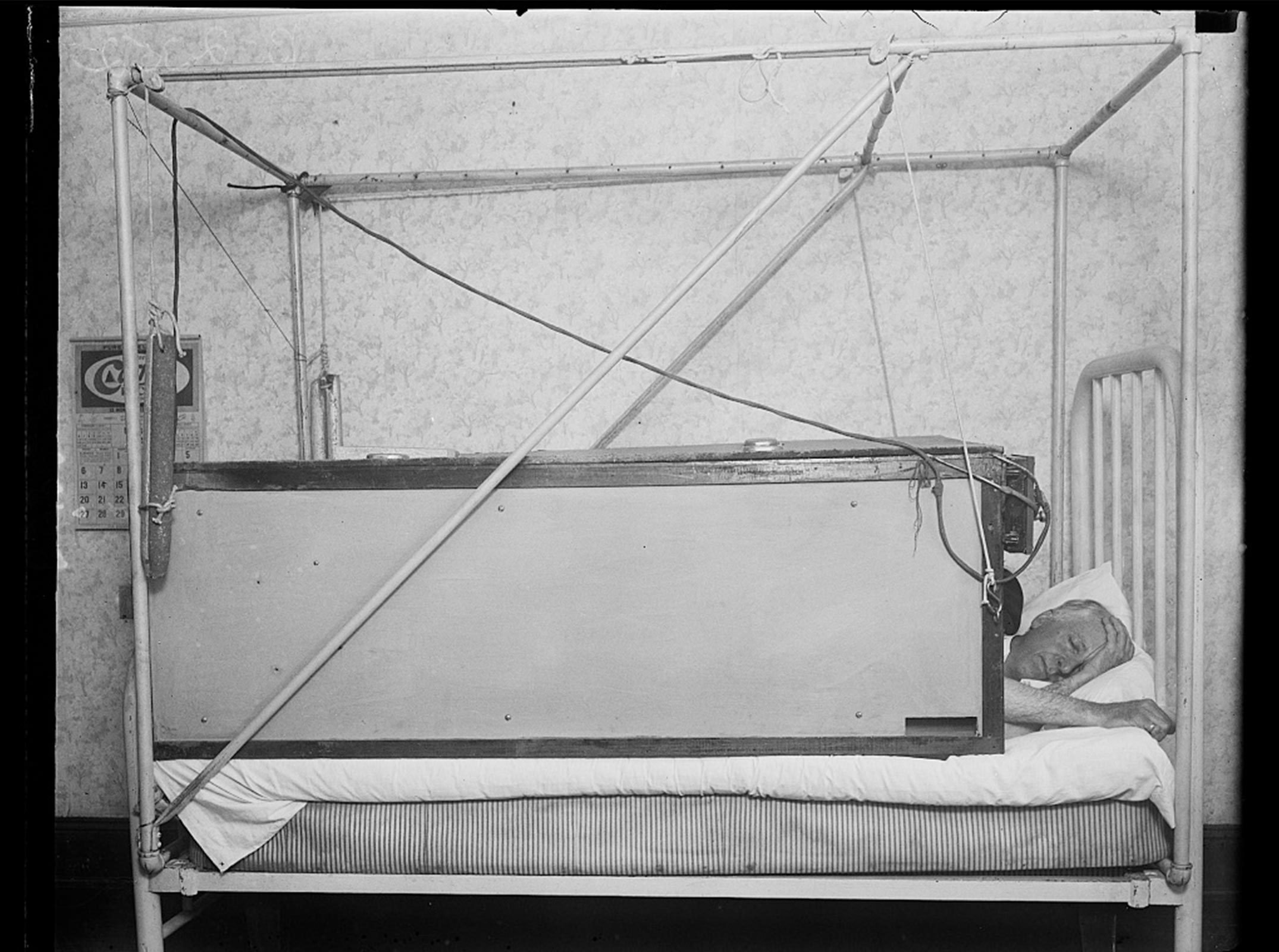 Man sleeps in vintage medical bed contraption, surrounded by metal frame and wires, in a sparsely decorated room with a visible calendar on the wall.