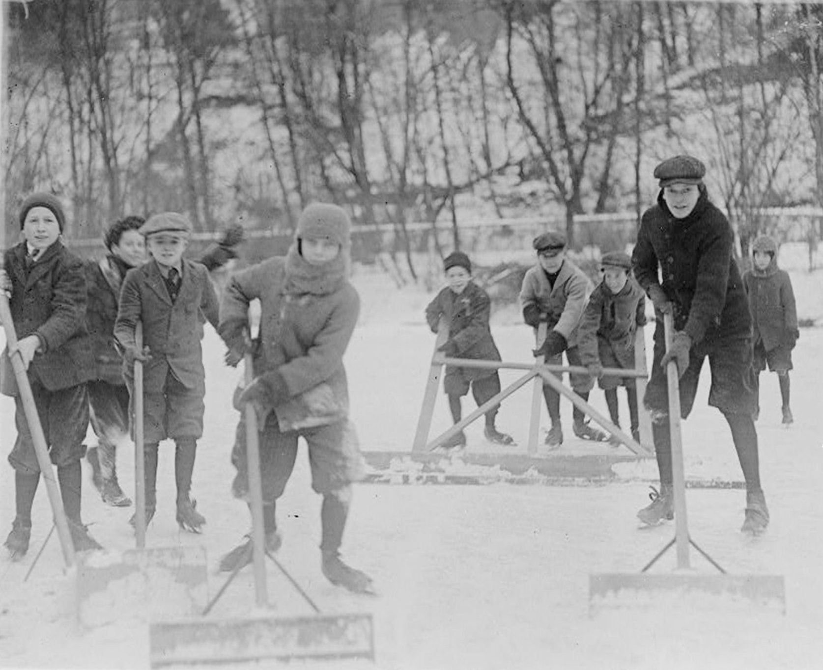 Children push snowplows, clearing a snowy patch on an outdoor ice rink, surrounded by leafless trees in a winter landscape.