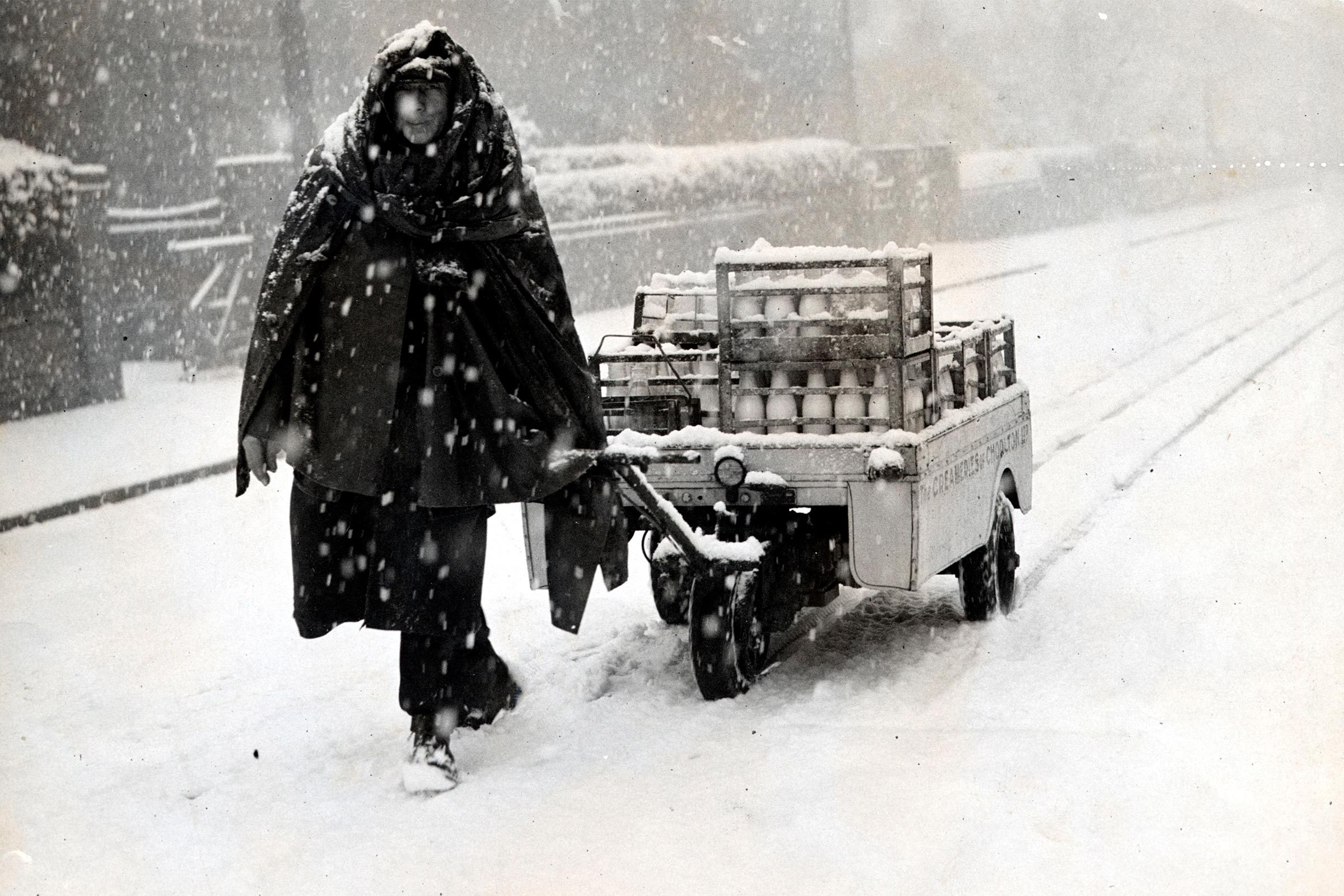 Person pulls cart loaded with milk bottles through heavy snow on a street, wearing thick, snow-covered cloak for warmth.