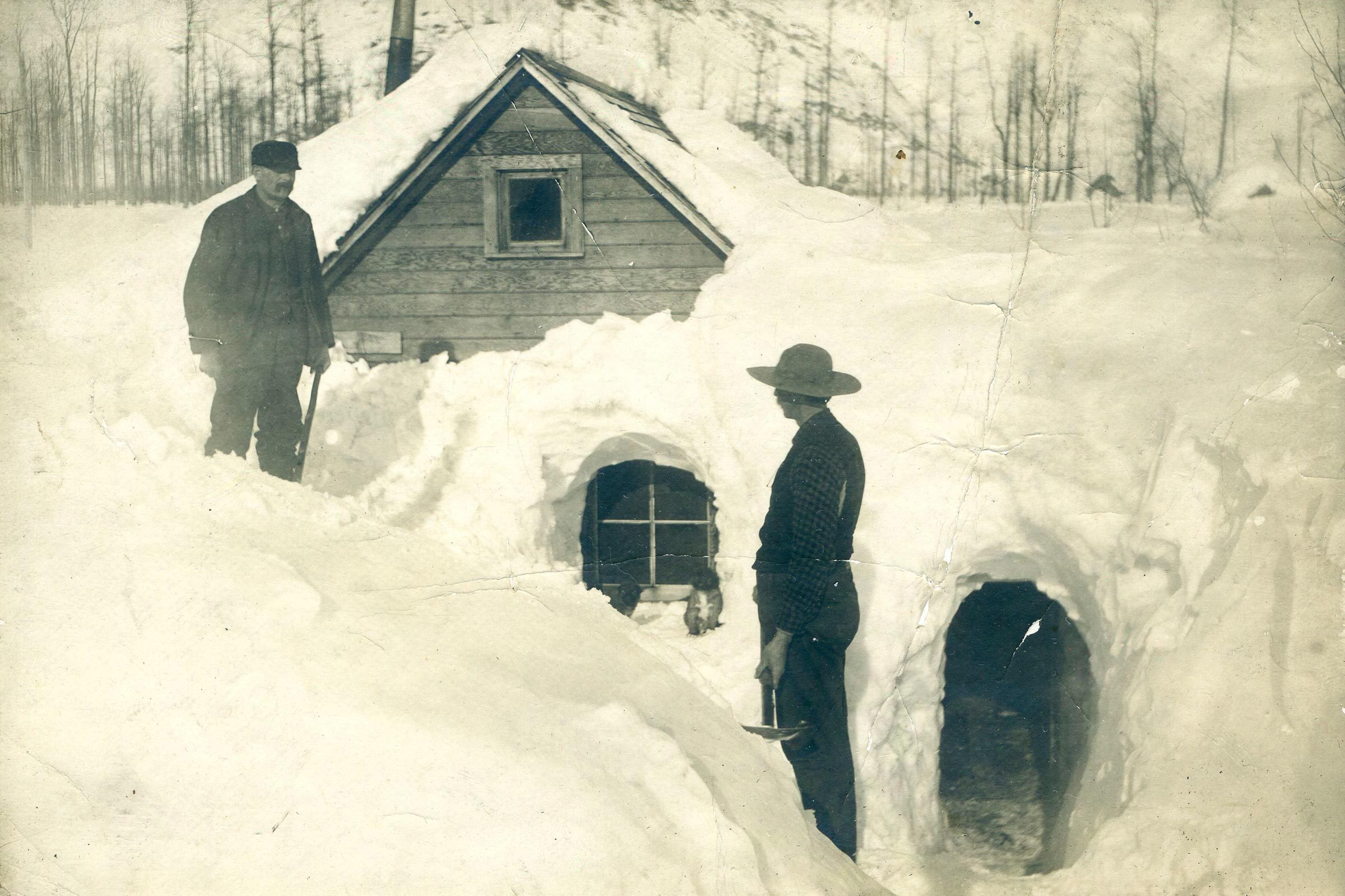 Two men stand by a snow-covered cabin, holding tools, surrounded by deep snow and bare trees.