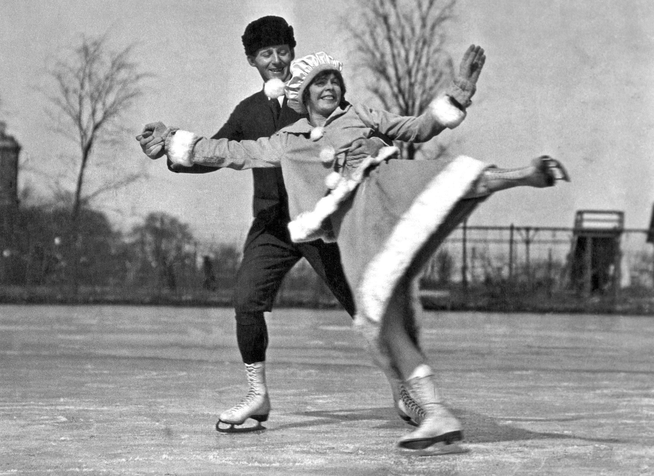 Two people ice skating gracefully, wearing winter outfits, outdoors on a frozen pond with bare trees in the background.