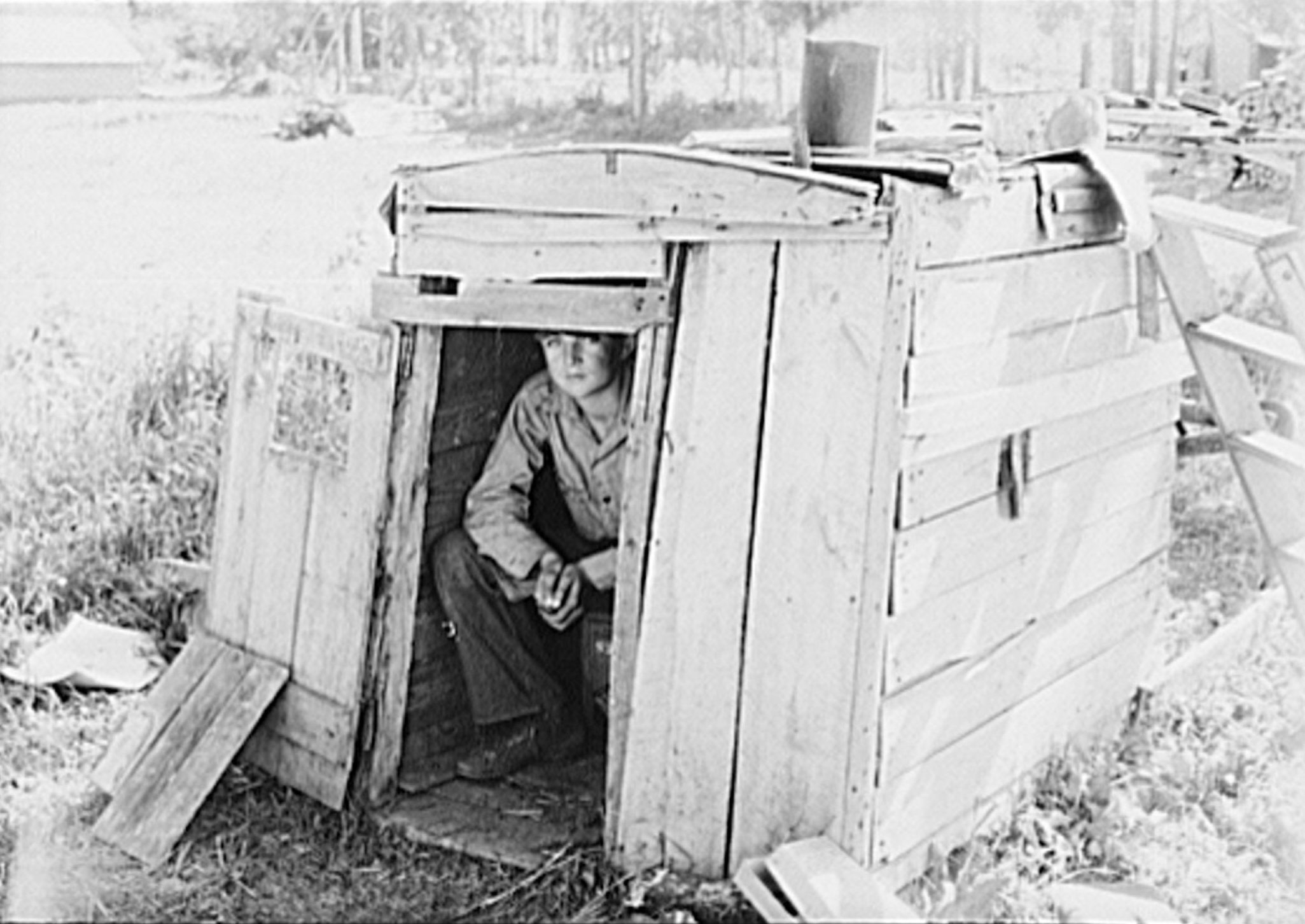 A person crouches inside a rustic wooden shed with an open door, surrounded by a grassy outdoor environment.