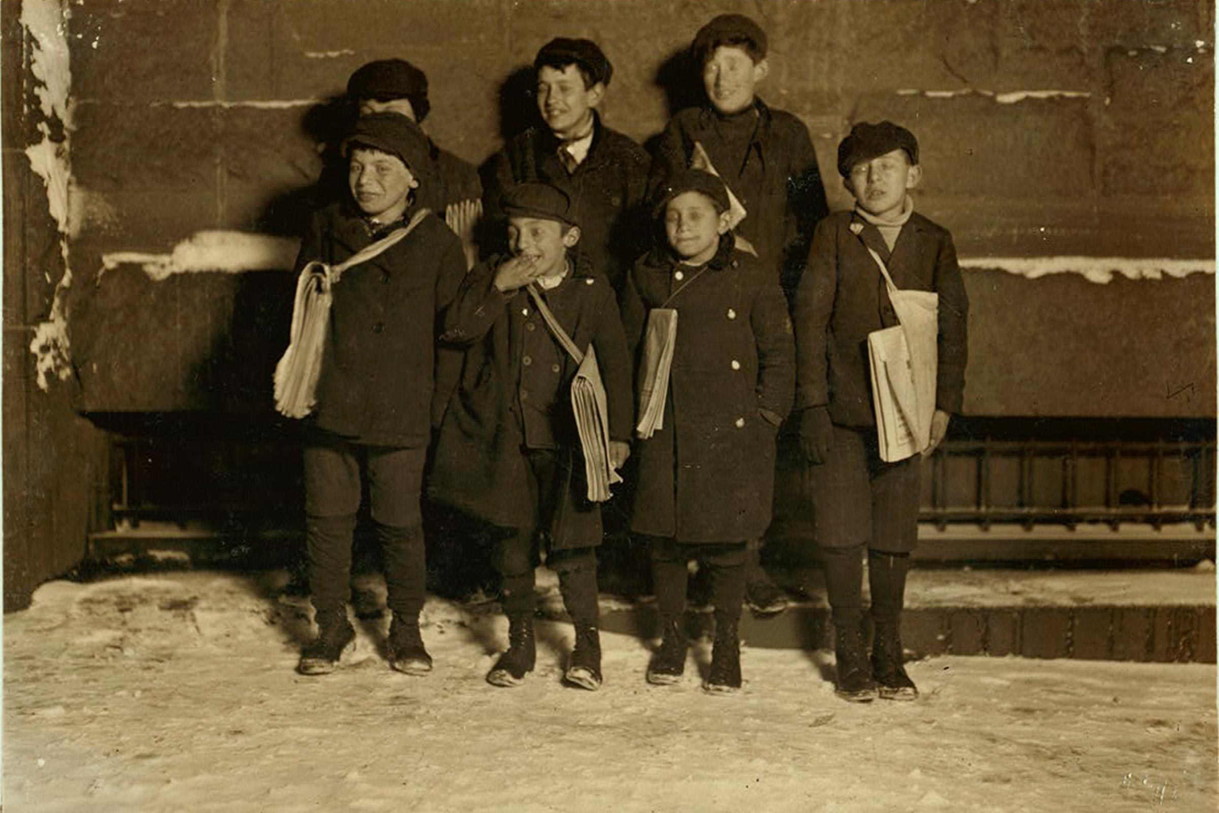 Children stand holding newspapers, wearing coats and hats, in a snowy urban setting against a stone wall.