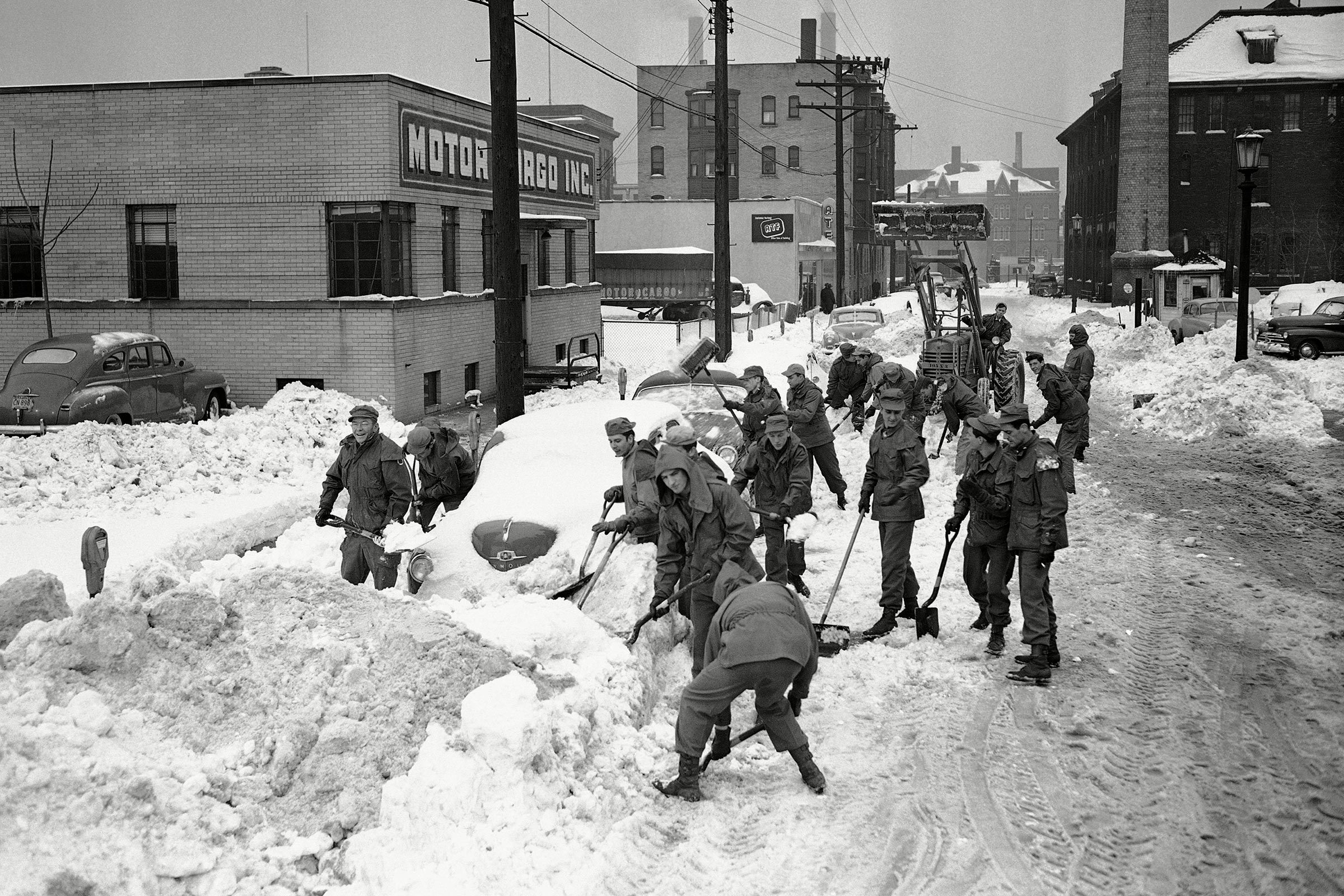 People shovel snow-covered street near brick building labeled "MOTOR CARGO INC." amidst parked cars; urban winter setting with utility poles.