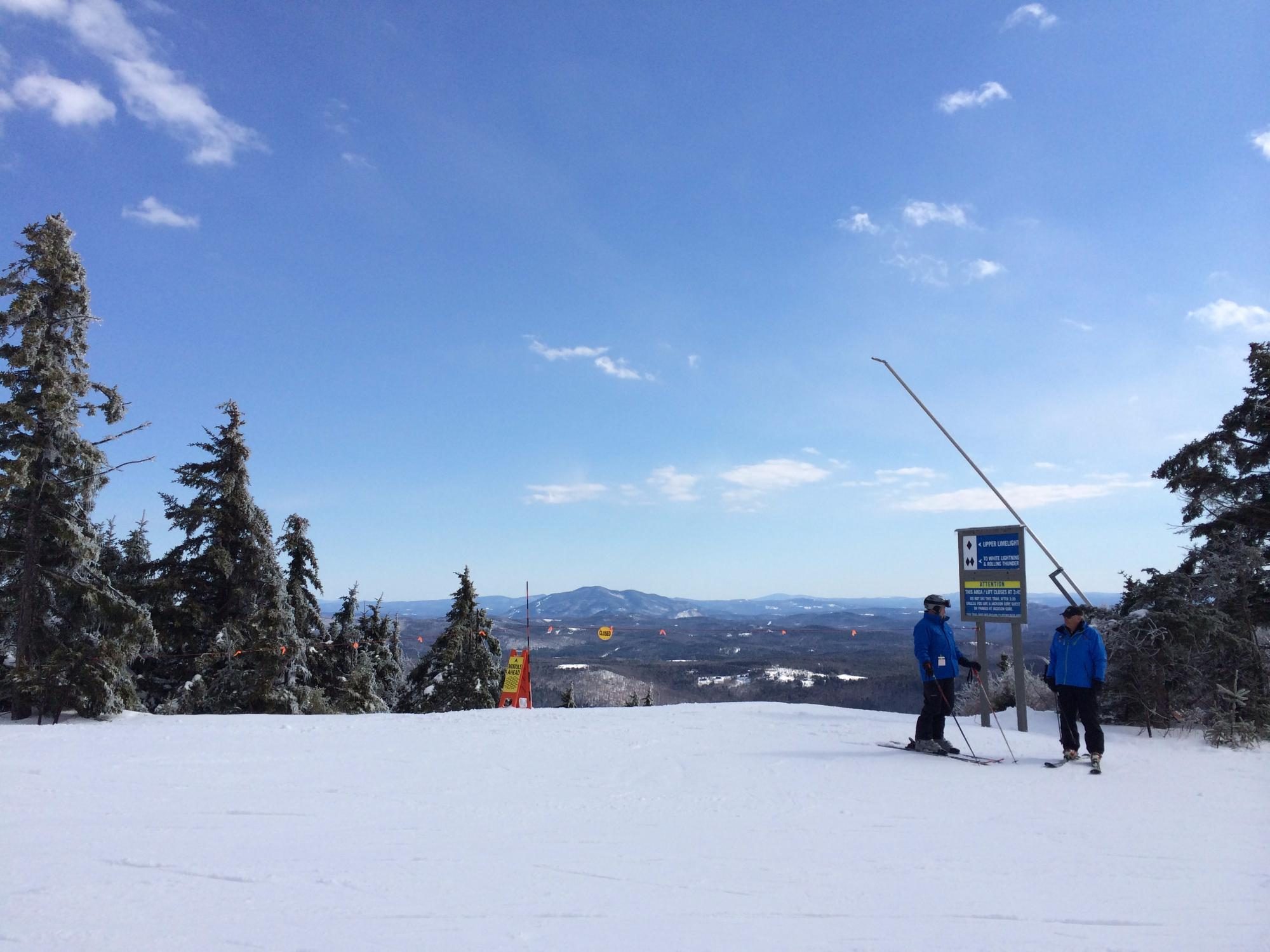 Skiers stand talking near a trail signpost against a snowy landscape with evergreen trees and distant mountains under a clear blue sky.