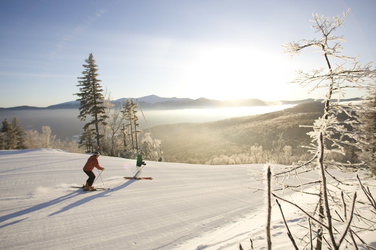 Skiers glide downhill on a snowy slope, surrounded by frosted trees and a bright sunrise over distant mountains.