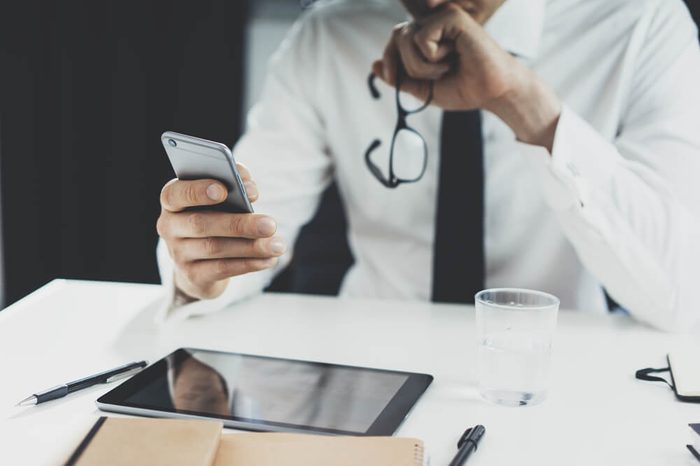 Close-up of businessman holding glasses and working on smartphone at his office, close-up of male hands using mobile phone, digital tablet and office tools on the desk