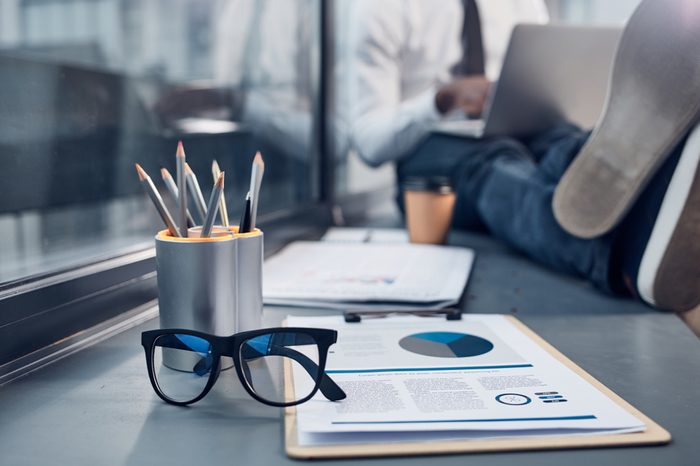 Necessary things. Close-up of document folder, trendy glasses and box of pencils are on windowsill with young businessman is sitting in background and typing on laptop