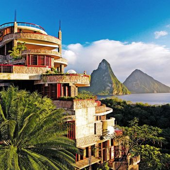 Modern, terraced building overlooks lush greenery and ocean, with two prominent peaks in the background under a clear blue sky.