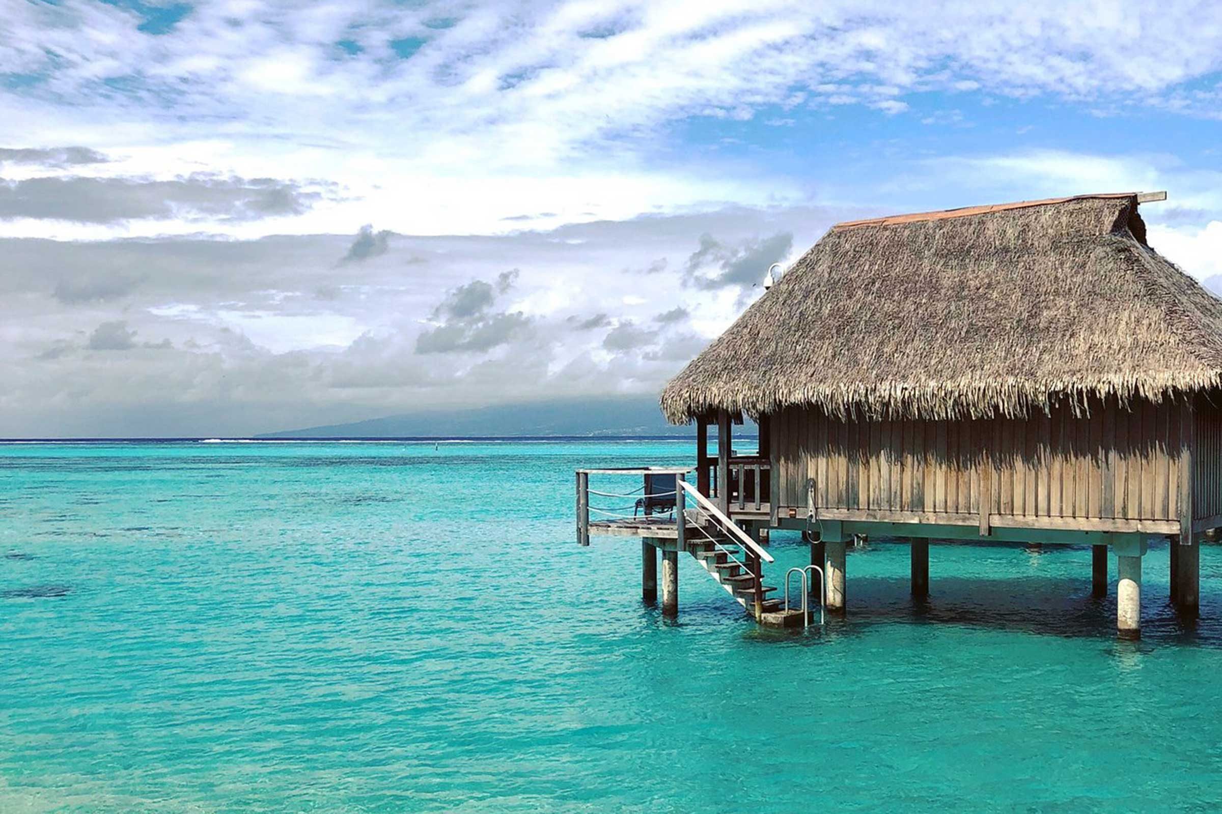 Thatched-roof hut stands on stilts over turquoise water, with a staircase descending into the sea, set under a partly cloudy sky.