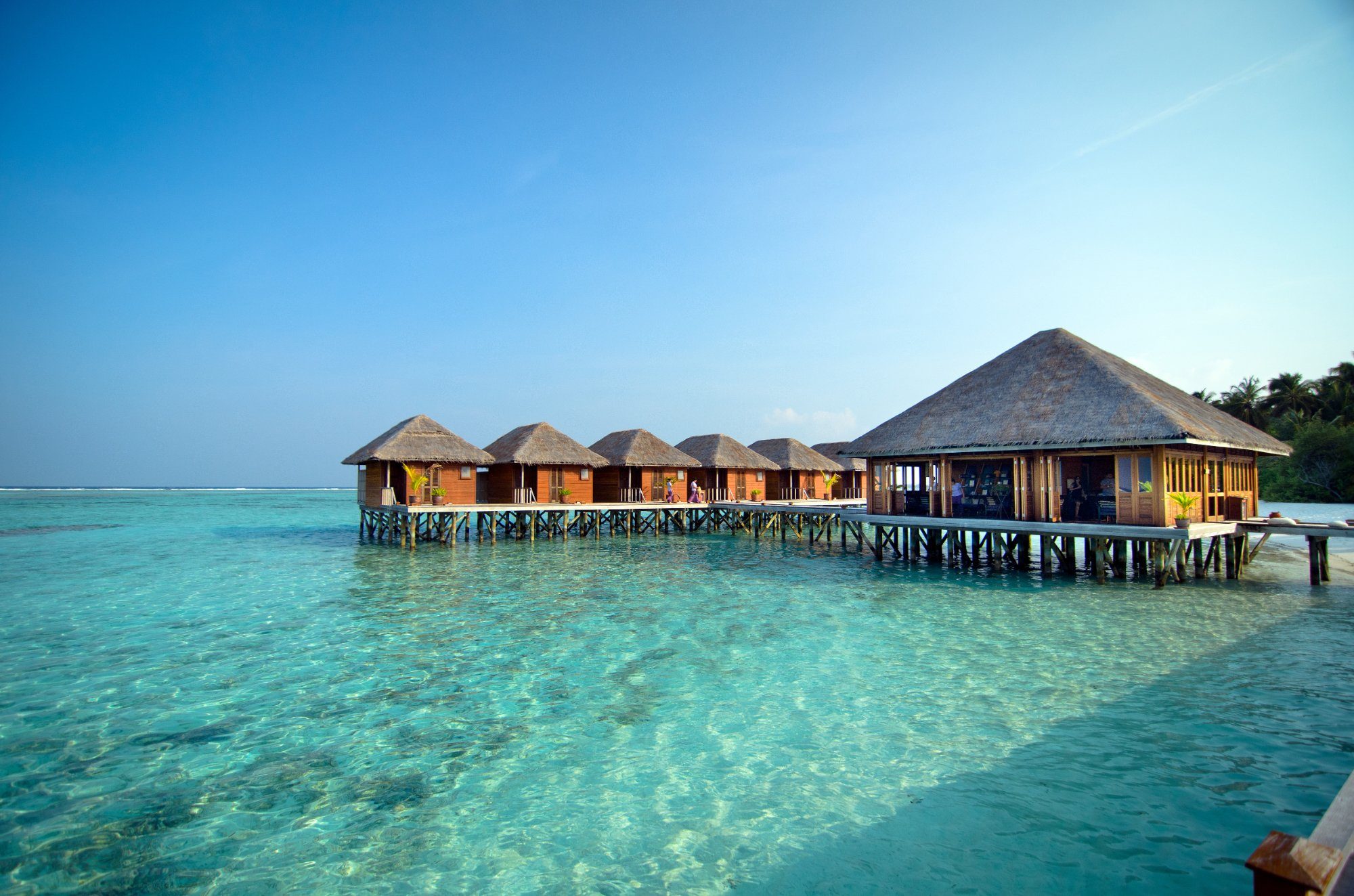 Wooden bungalows stand elevated above turquoise water, connected by a boardwalk, under a clear blue sky.