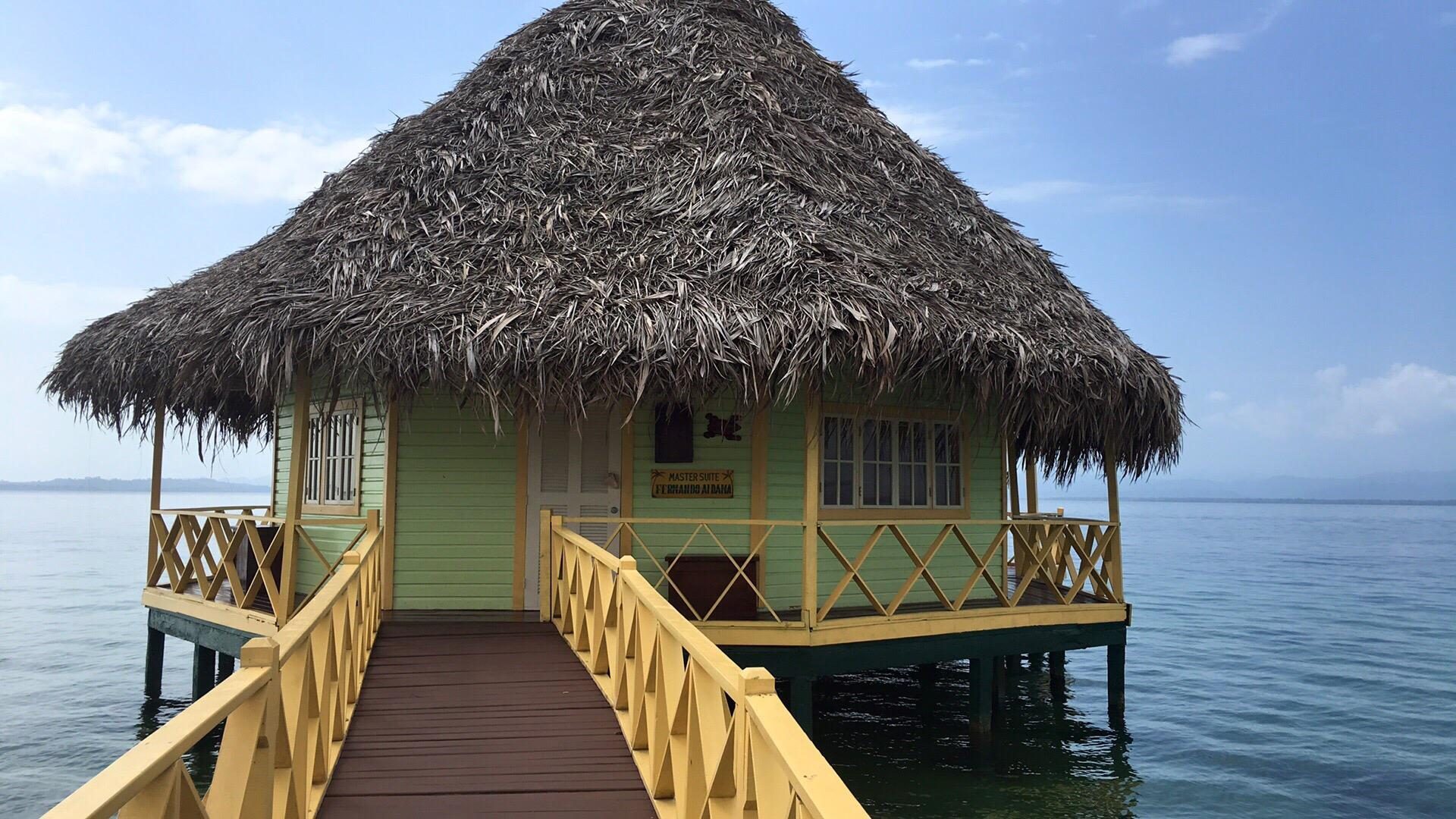 Thatched-roof bungalow stands on stilts over calm water, connected by a wooden walkway, under a partly cloudy sky. Sign reads: "Master Suite JF Giraud Audeaga."
