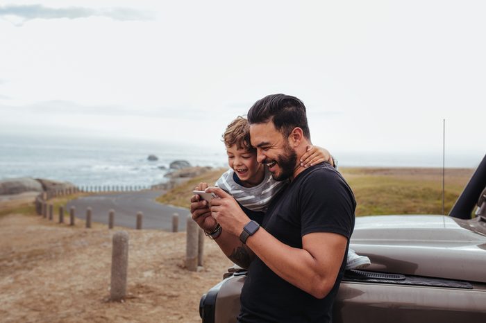 Outdoor shot of happy father and son in front of the car using mobile phone and laughing. Young man and little boy watching something on smart phone and enjoying while on road trip.