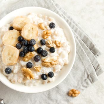 Bowl of oatmeal topped with banana slices, blueberries, and walnuts on a gray napkin with a spoon nearby on a light surface.