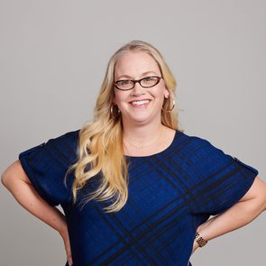 A person smiles confidently, hands on hips, wearing a blue patterned shirt and glasses, against a plain gray background.