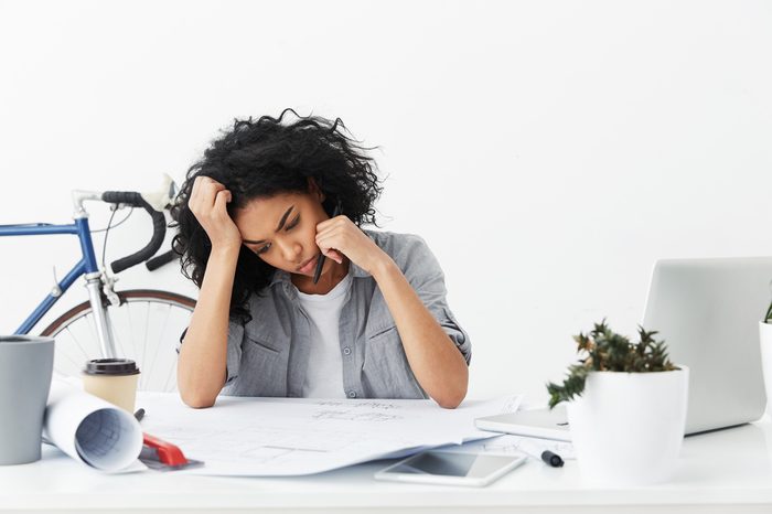 Portrait of bored and tired Afro American female architect working on architectural plan unwillingly, feeling indifference, sitting at white office desk with blueprints and electronic devices