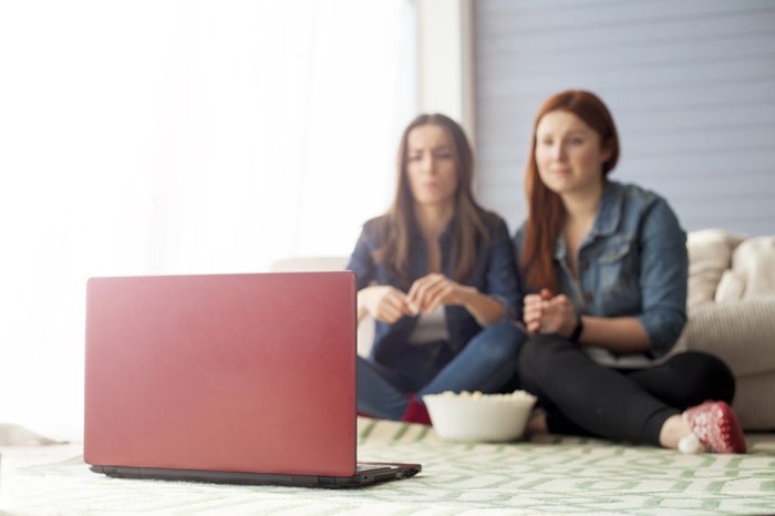 Two girl friends sitting on the floor near the couch in front of them there is a computer on which they watch a movie and eat popcorn. Funny friends relaxing together.
