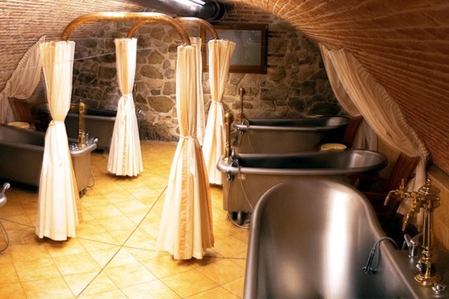 Bathtubs are arranged under an arched, brick ceiling with surrounding privacy curtains, in a stone-walled room on a tiled floor.