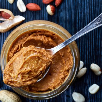 Jar and spoon of peanut butter and peanuts on dark wooden background from top view