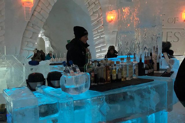 Bottles sit on an ice bar, tended by a person wearing winter clothes, inside an ice structure with blue lighting and ice walls.