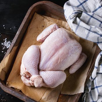 Raw organic uncooked whole chicken with salt and pepper on backing paper in old oven tray with white kitchen towel over black burnt wooden background. Top view with space.