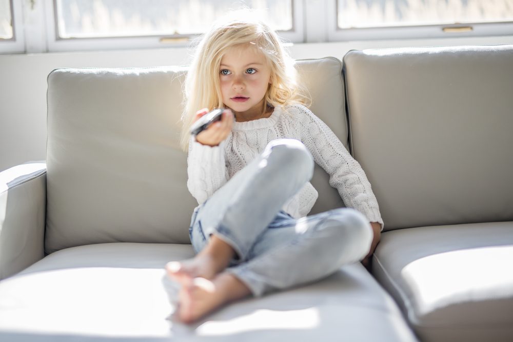 A 4 years old child watching tv laying down on the sofa at home alone