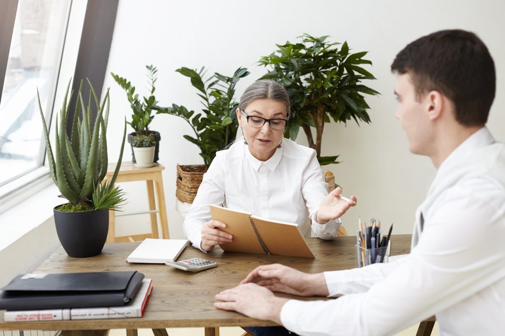 Confident attractive woman CEO in her fifties holding copybook while posing questions about job qualification, experience and skills during interview with brunette male applicant. Film effect