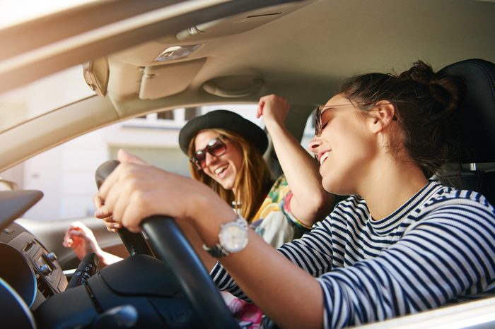 Two trendy attractive young woman singing along to the music as they drive along in the car through town viewed through the open side window