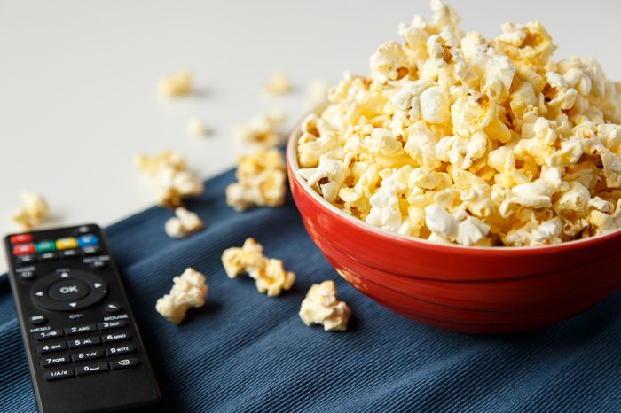 Popcorn in red bowl with tv remote on white table, selective focus.
