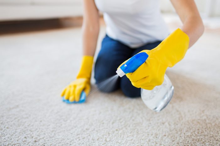 people, housework and housekeeping concept - close up of woman in rubber gloves with cloth and derergent spray cleaning carpet at home
