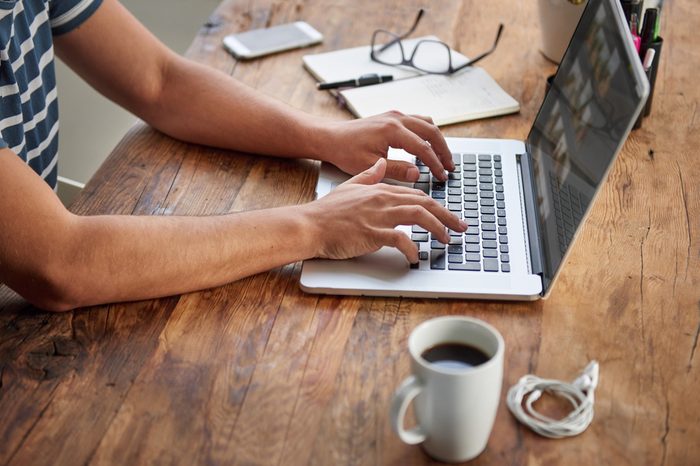 Cropped shot of a man's hands typing on a laptop that is on a wooden desk with a mug of coffee alongside