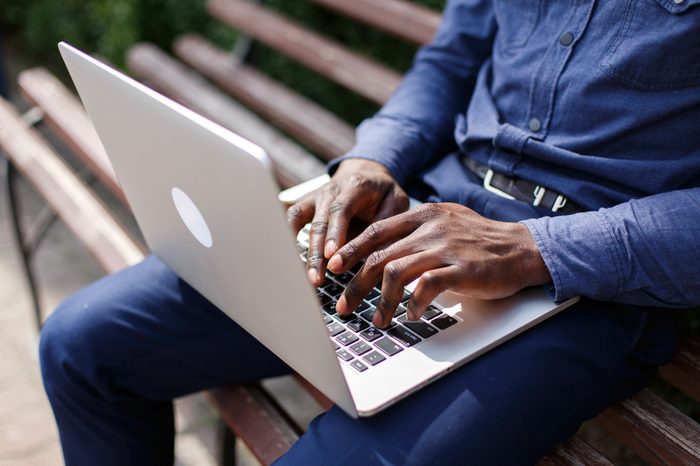 Hands of African American man typing something on the laptop while he sits on the bench outside