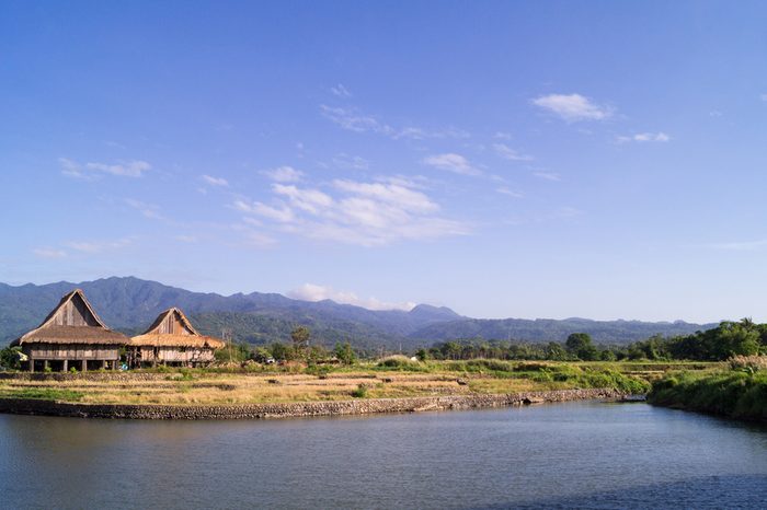 A scenic of view of the local landscape in Bagac, Bataan, Philippines. Two wooden houses stand along a river. Mountains surround the area.