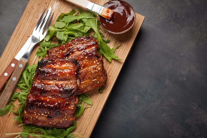 Closeup of pork ribs grilled with BBQ sauce and caramelized in honey on a bed of arugula. Tasty snack to beer on a wooden Board for filing on dark concrete background. Top view with copy space.