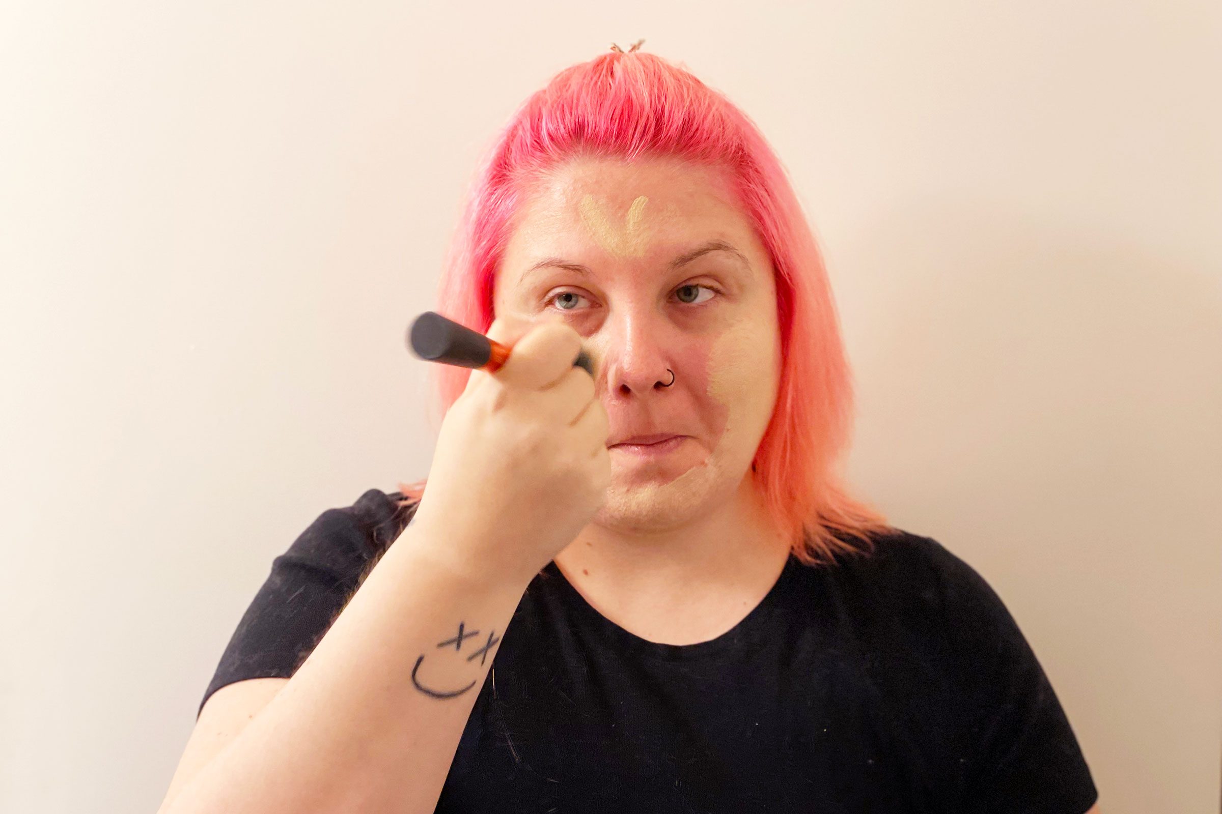 Person applying makeup with brush, wearing black shirt, pink hair, against plain background. Arm tattoo: smiling face with X for eyes.