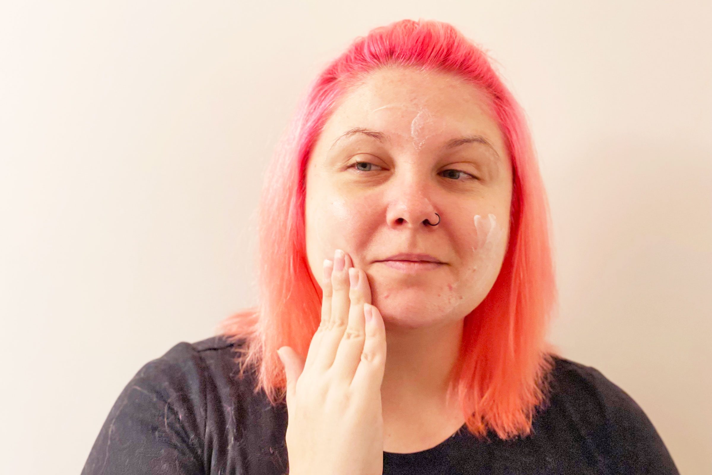 Person with pink hair applying face cream, standing indoors against a plain background.