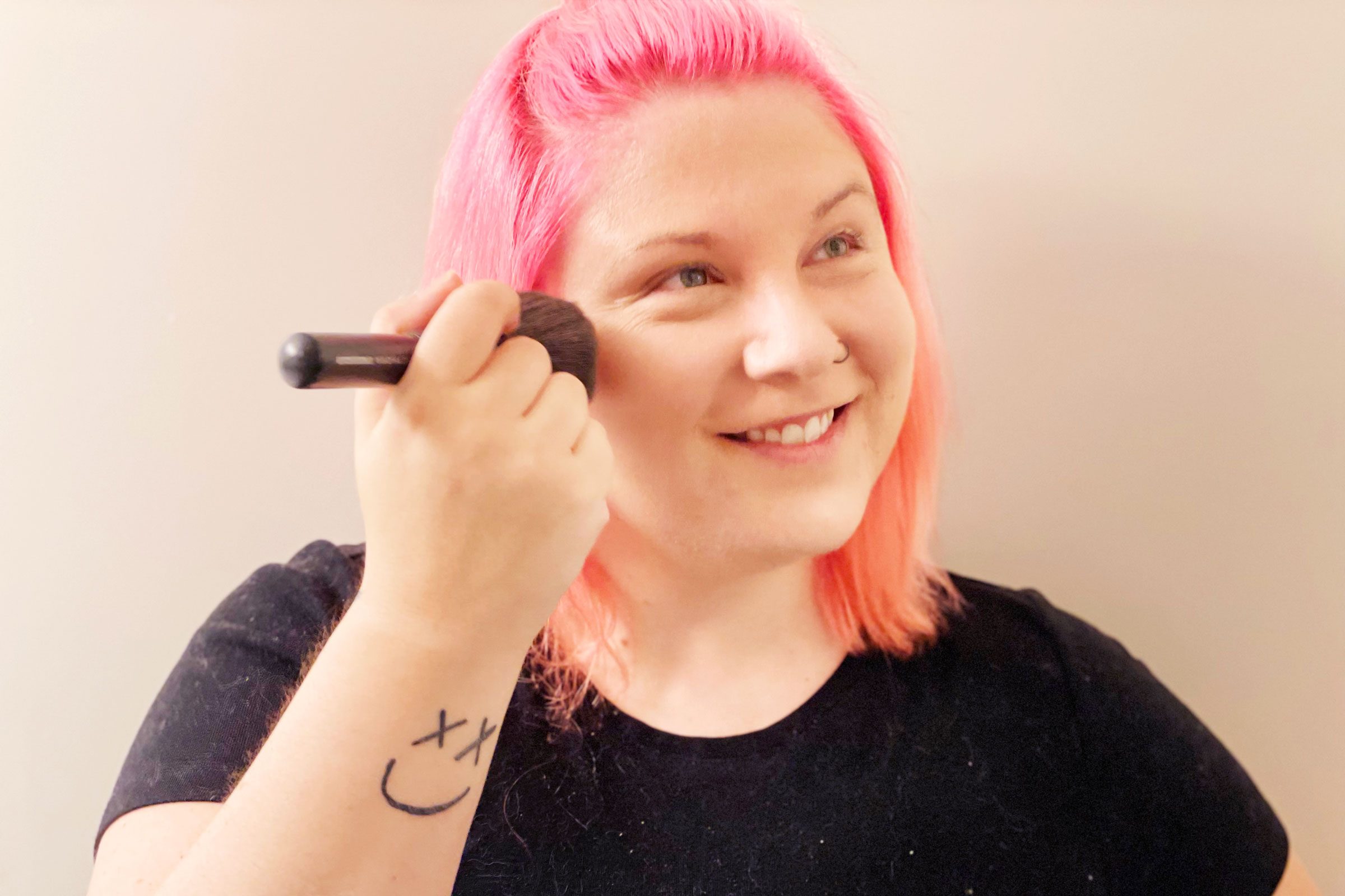 Person uses makeup brush, smiling with pink hair, wearing a black shirt indoors. Smiley face tattoo on forearm.