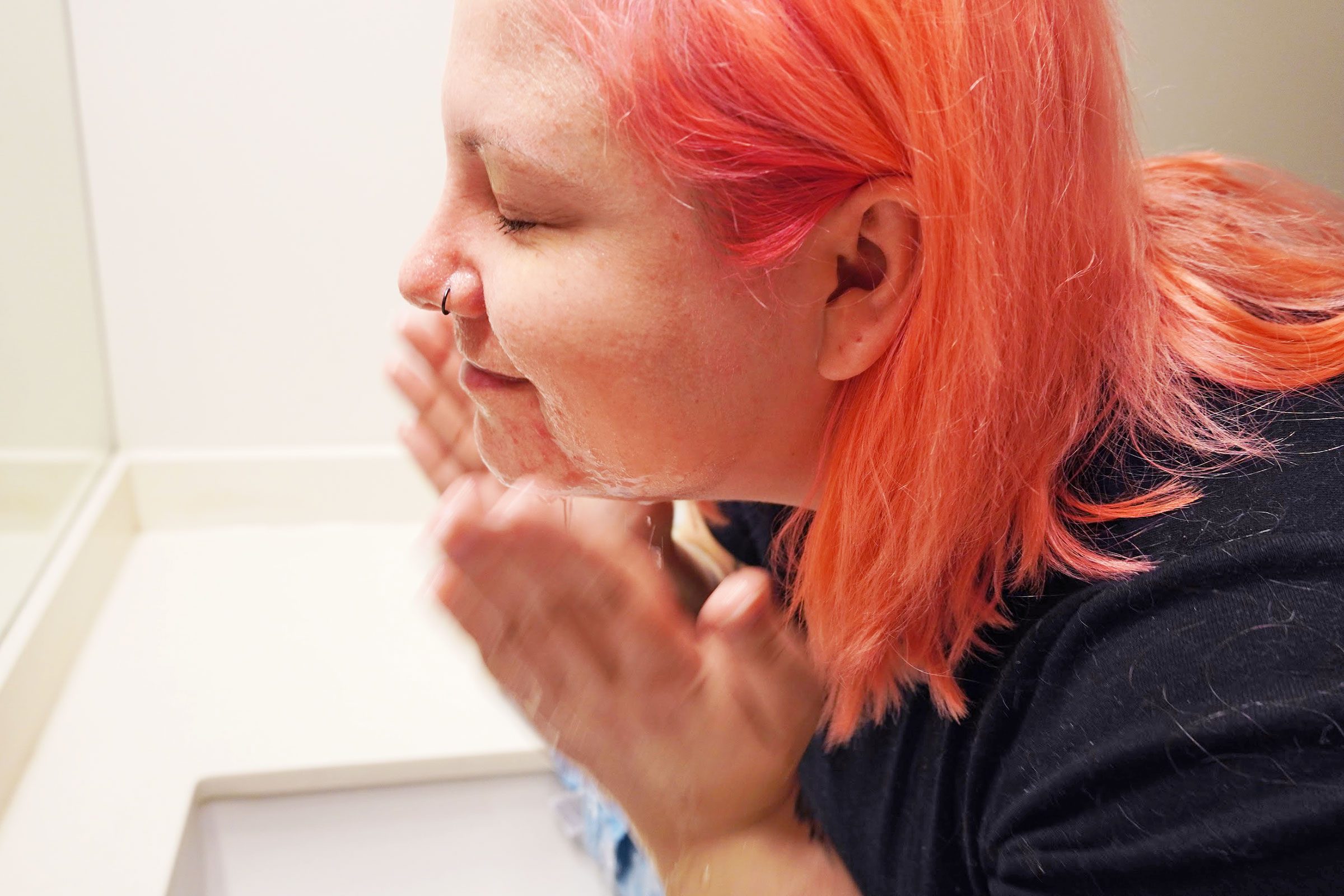 Person washing face, lathering soap, in a bathroom with a white sink and mirror.