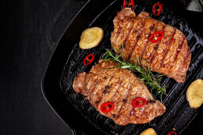 Grilled pork steak in grill pan with rosemary, pepper chilli and ginger on wooden board. Top view. Copy space.