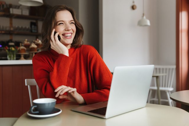 Image of happy caucasian woman 20s laughing and talking on smartphone while using laptop in cafe