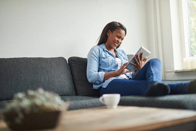 Young African woman laughing while relaxing on her living room sofa browsing online with a digital tablet
